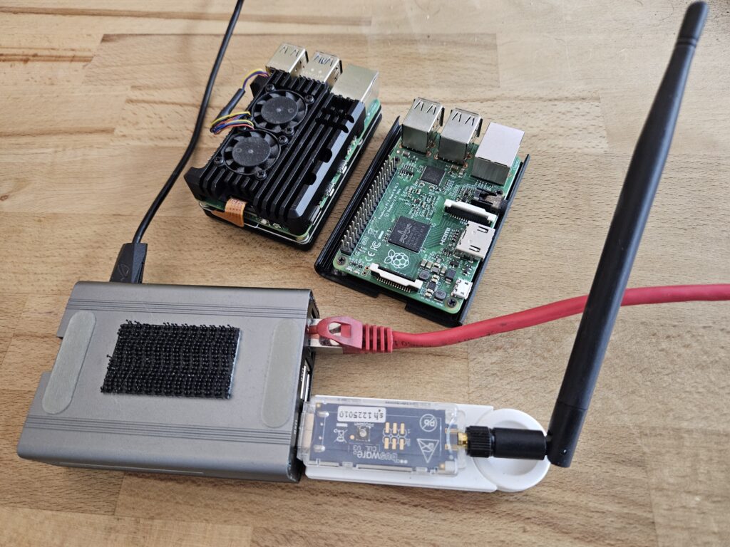Auf einem hölzernen Tisch liegen mehrere Einplatinencomputer und Zubehörteile. Links vorne befindet sich ein rechteckiges, graues Gehäuse mit Klettfläche, aus dem ein rotes Netzwerkkabel führt. Daran angeschlossen ist ein USB-Stick mit transparenter Hülle und aufgeschraubter schwarzer Antenne. Dahinter stehen zwei Raspberry Pi Platinen: eine mit schwarzem Kühlkörper und zwei kleinen Lüftern, die andere mit frei sichtbarer Platine, Ports und dem Raspberry-Logo.