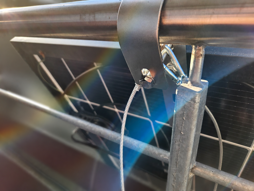 Nahaufnahme der Befestigung eines Solarmoduls am Balkongeländer. Zusätzlich zur eigentlichen Halterung ist ein Sicherungsseil angebracht, welches im Falle eines mechanischen Versagens einen Absturz des Moduls verhindern soll.
